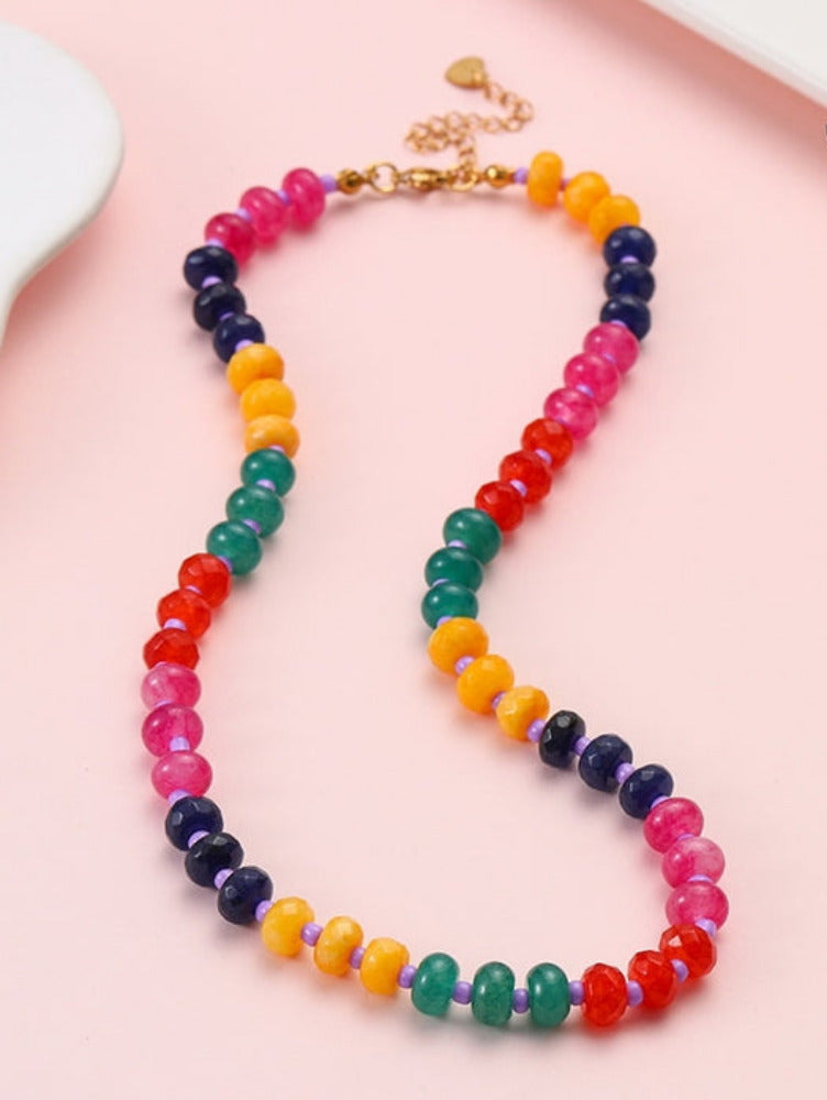 A colorful beaded necklace with alternating sections of pink, orange, yellow, green, red, and dark purple beads, arranged in an S-shape on a light pink background.