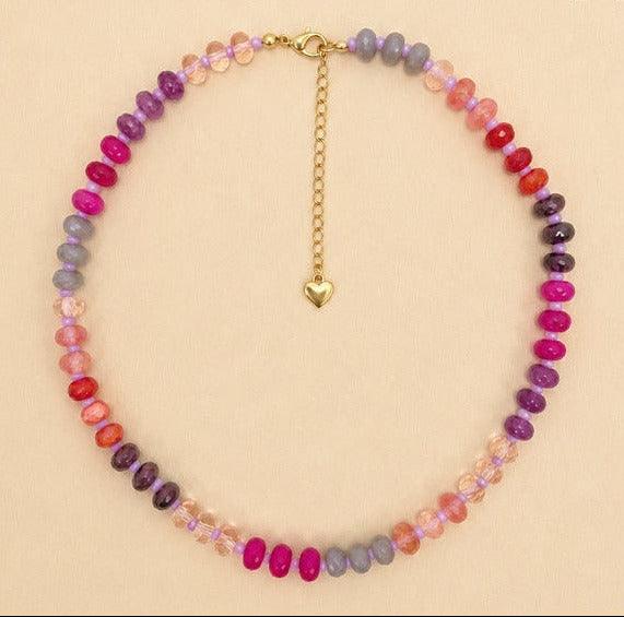 A beaded choker necklace with alternating rows of faceted pink, purple, and gray beads, accented by a gold clasp, extender chain, and small gold heart charm. The necklace is displayed on a solid beige background.