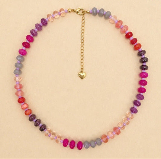A beaded choker necklace with alternating rows of faceted pink, purple, and gray beads, accented by a gold clasp, extender chain, and small gold heart charm. The necklace is displayed on a solid beige background.
