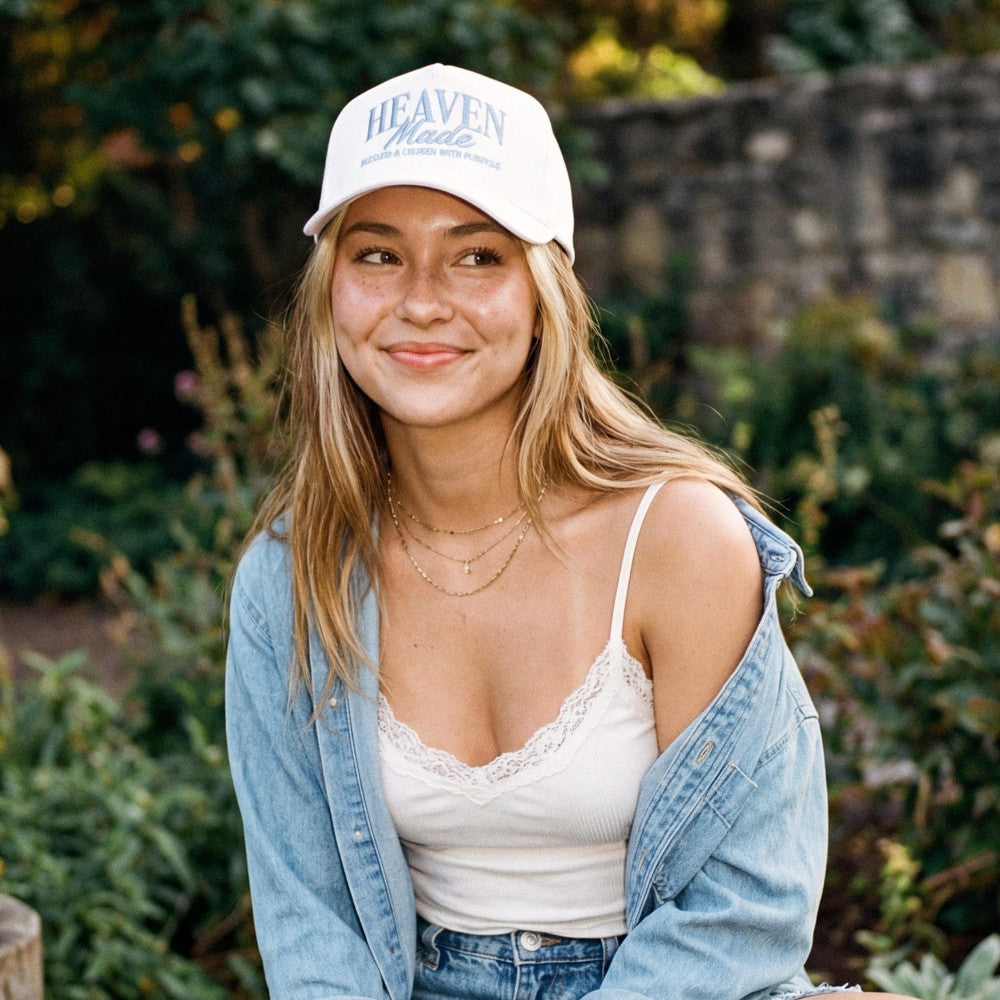 A young woman wearing a white 'Heaven Made' baseball cap, a white lace-trim camisole, an open light blue denim shirt, and jeans, set against a blurred outdoor garden background.