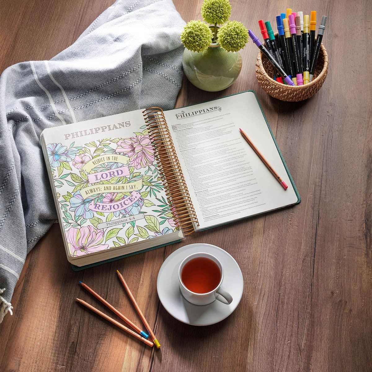 An open spiral-bound Bible lies on a wooden table, surrounded by a gray throw blanket, pencils, a cup of tea, and a small vase with green pom-pom flowers. A basket of colored pens or markers sits in the upper right.