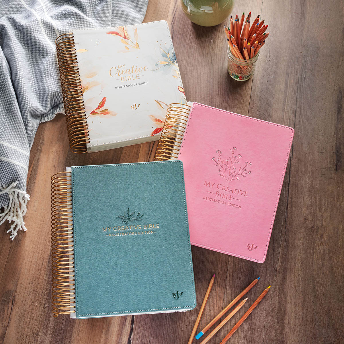 Three 'My Creative Bible' notebooks in blue, pink, and white on a wooden surface with pencils.