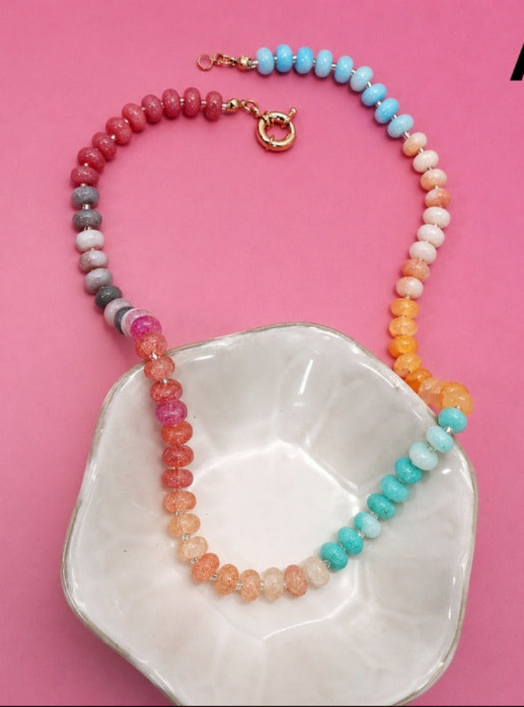 A beaded necklace with multiple strands of round gemstone beads in pink, orange, yellow, blue, and gray, draped over a white ceramic dish on a solid pink background.